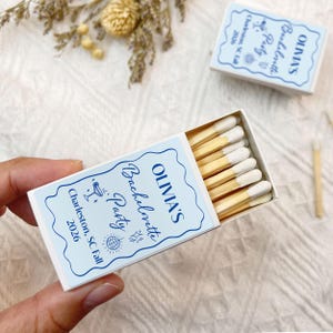 May include: White matchbox with blue and white text that reads "OLIVIA'S Bachelorette Party Charleston, SC Fall 2026". The matchbox is open and contains wooden matches with white tips.