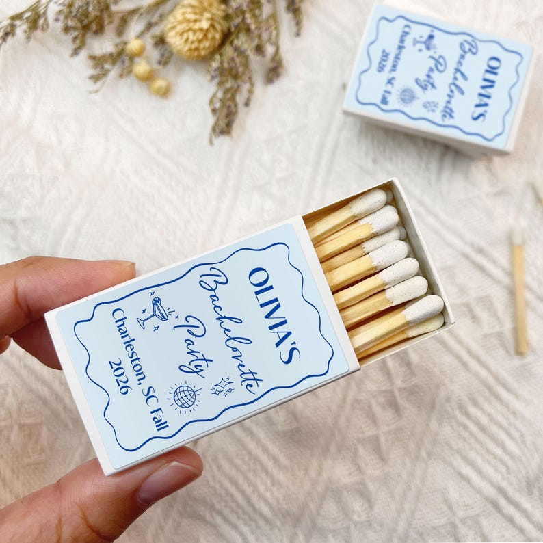 May include: White matchbox with blue and white text that reads "OLIVIA'S Bachelorette Party Charleston, SC Fall 2026". The matchbox is open and contains wooden matches with white tips.
