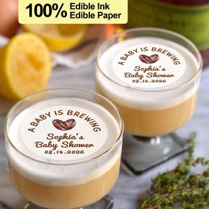 Può includere: Due bicchieri trasparenti pieni di una bevanda a strati, sormontati da schiuma bianca. Ogni bicchiere ha una carta commestibile circolare con il testo "A BABY IS BREWING" e "Sophia's Baby Shower 08.14.2026". L'immagine include anche il testo "100% Edible Ink Edible Paper".