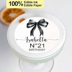K&ouml;nnte beinhalten: Runder essbarer Kuchentopper in Wei&szlig; mit einer schwarzen Schleife und dem Text "Isabella N&deg;21 GEBURTSTAG".