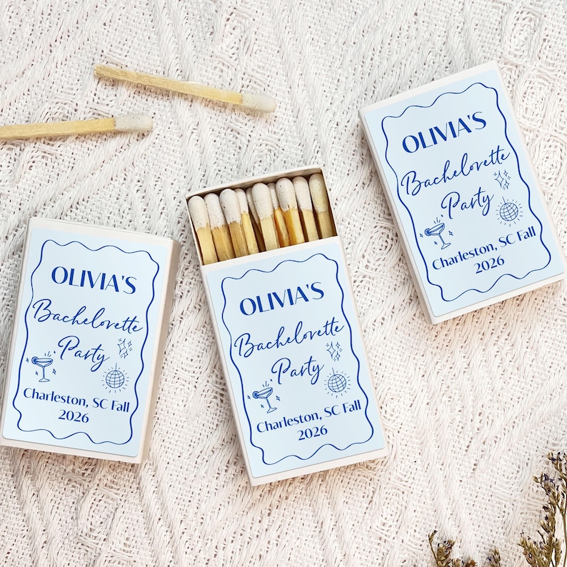 May include: Three white matchboxes with blue and white labels. The labels have the text "OLIVIA'S Bachelorette Party" with a cocktail glass and disco ball icon. The text also includes "Charleston, SC Fall 2026".
