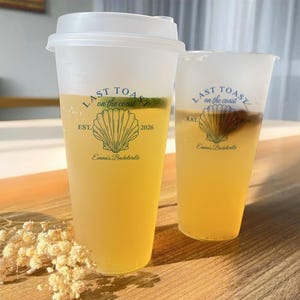 K&ouml;nnte beinhalten: Zwei durchsichtige Plastikbecher mit Deckeln, gef&uuml;llt mit einem hellgelben Getr&auml;nk. Die Becher haben ein Design mit dem Text "Last Toast on the Coast", einer Muschel und dem Text "Emma's Bachelorette" und "EST. 2026".