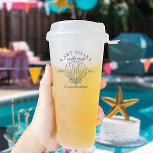 K&ouml;nnte beinhalten: Ein durchsichtiger Plastikbecher mit wei&szlig;em Deckel und einem blauen und wei&szlig;en Design, auf dem "Last Toast on the Coast est. 2026" mit einer Muschelgrafik und dem Text "Emma's Bachelorette" steht.