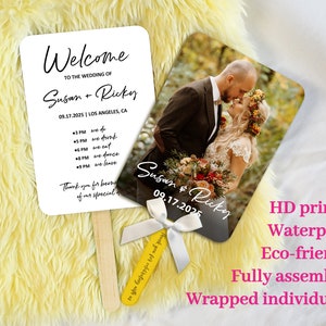 Può includere: Un ventaglio da matrimonio bianco con testo nero che dice "Benvenuti al matrimonio di Susan + Ricky 09.17.2025 | Los Angeles, CA" e un programma di eventi. Il ventaglio ha un bastoncino di legno e un nastro bianco. Il ventaglio è accanto a un ventaglio simile con una foto di una sposa e uno sposo che si baciano. Il testo sul ventaglio fotografico dice "Susan & Ricky 09.17.2025". Il testo sotto i ventagli dice "Stampa HD Impermeabile Ecologico Completamente assemblato Confezionato singolarmente".