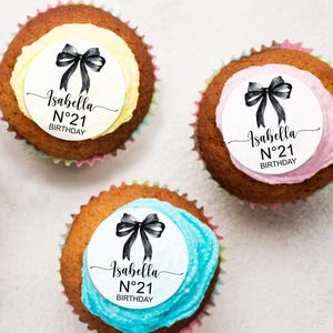 K&ouml;nnte beinhalten: Drei Cupcakes mit wei&szlig;em Frosting und schwarzen Schleifen. Jeder Cupcake hat einen runden wei&szlig;en Aufkleber mit dem Text "Isabella N&deg;21 BIRTHDAY" in Schwarz.