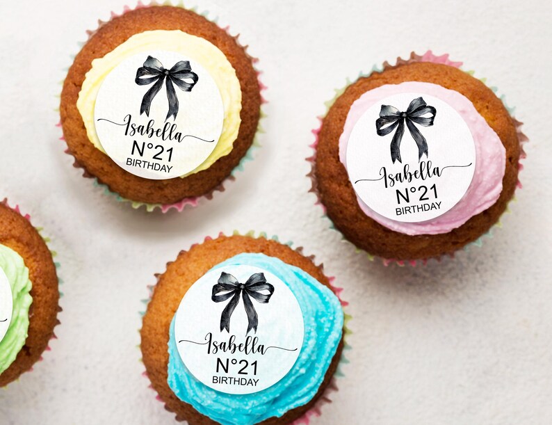 K&ouml;nnte beinhalten: Drei Cupcakes mit wei&szlig;em Frosting und schwarzen Schleifen. Jeder Cupcake hat einen runden wei&szlig;en Aufkleber mit dem Text "Isabella N&deg;21 BIRTHDAY" in Schwarz.