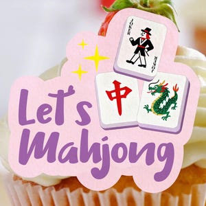 Könnte beinhalten: Ein Cupcake mit einer rosa Dekoration mit der Aufschrift "Let's Mahjong". Die Dekoration zeigt eine Joker-Karte und zwei Mahjong-Steine, einer mit einem roten Zeichen und der andere mit einem grünen Drachen. Gelbe Sterne sind ebenfalls vorhanden.