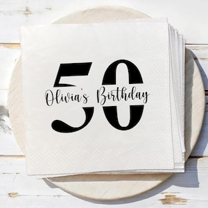 Può includere: Una pila di tovaglioli di carta bianchi con un "50" nero e la scritta "Olivia's Birthday" in corsivo. I tovaglioli sono su un vassoio di legno, perfetti per una festa di compleanno per i 50 anni.