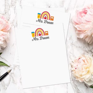 Può includere: Due blocchi note bianchi con un design a forma di arcobaleno, una mela, un libro e una tazza di caffè. Il testo "Mrs. Brown" è stampato su ogni blocco note.