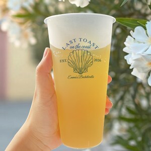 K&ouml;nnte beinhalten: Ein durchsichtiger Plastikbecher mit einem gelben Getr&auml;nk. Der Becher hat ein wei&szlig;es Design mit dem Text "Last Toast on the coast" und einer Muschelgrafik. Der Text "Est. 2026" und "Emma's Bachelorette" ist ebenfalls auf dem Becher aufgedruckt.