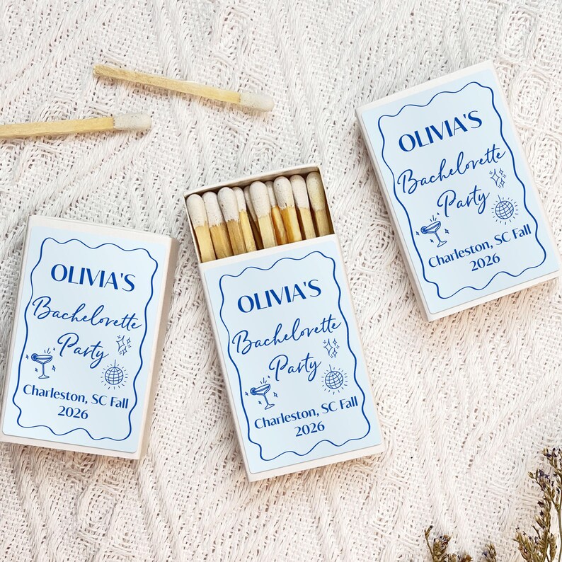 May include: Three white matchboxes with blue and white labels. The labels have the text "OLIVIA'S Bachelorette Party" with a cocktail glass and disco ball icon. The text also includes "Charleston, SC Fall 2026".