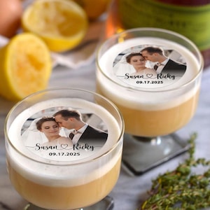 May include: Two clear glass cocktail glasses containing a white frothy drink. Each glass has a round sticker with a photo of a bride and groom, the text "Susan ♥ Ricky" and the date "09.17.2025".