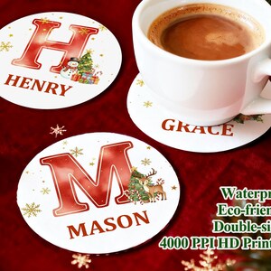 May include: White Christmas coasters with red letters and names, including "HENRY", "MASON", and "GRACE". Each coaster features a festive design with snowflakes, a Christmas tree, and a snowman. A white mug of coffee sits on one coaster.
