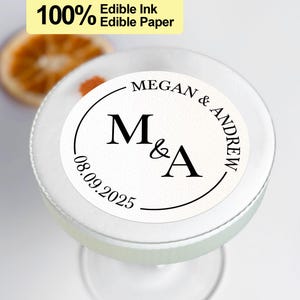 May include: White round edible paper sticker with black text. The text reads "MEGAN & ANDREW" in a circle around the letters "M&A". The date "08.09.2025" is below the letters.