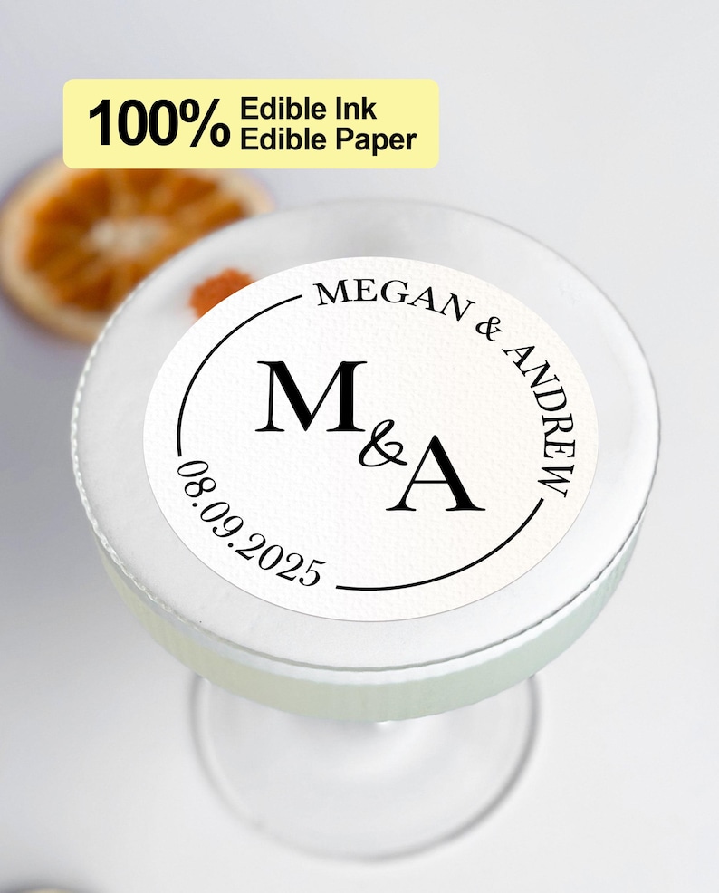 May include: White round edible paper sticker with black text. The text reads "MEGAN & ANDREW" in a circle around the letters "M&A". The date "08.09.2025" is below the letters.