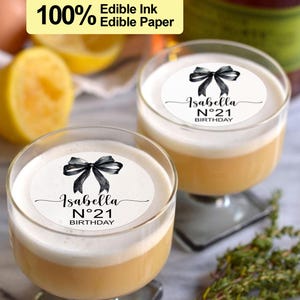K&ouml;nnte beinhalten: Zwei klare Glasbecher mit wei&szlig;em Hintergrund und schwarzem Text, der "Isabella N&deg;21 BIRTHDAY" lautet, mit einer schwarzen Aquarell-Schleife oben. Die Becher sind mit einer hellgelben Fl&uuml;ssigkeit gef&uuml;llt.