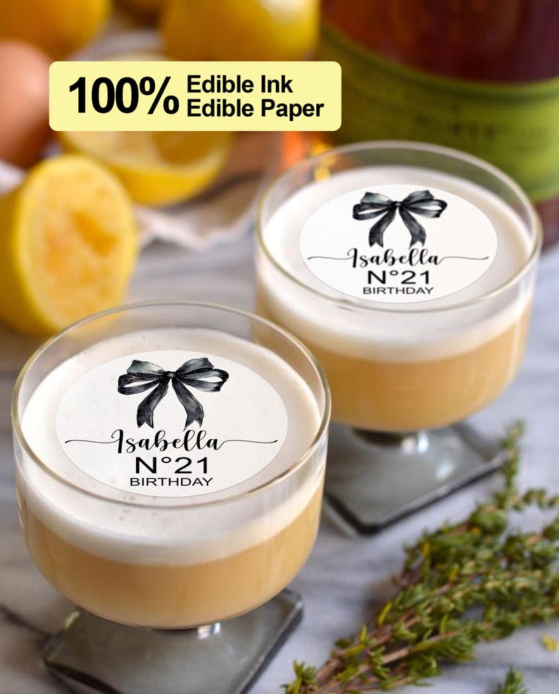 K&ouml;nnte beinhalten: Zwei klare Glasbecher mit wei&szlig;em Hintergrund und schwarzem Text, der "Isabella N&deg;21 BIRTHDAY" lautet, mit einer schwarzen Aquarell-Schleife oben. Die Becher sind mit einer hellgelben Fl&uuml;ssigkeit gef&uuml;llt.