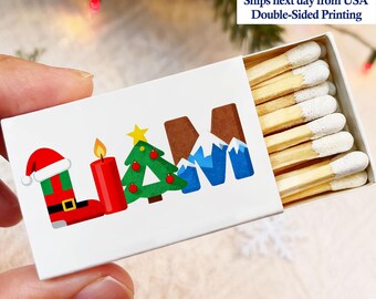 May include: A white matchbox with the name "LIAM" in a festive Christmas design. The letters are decorated with a Santa boot, a candle, a Christmas tree, and mountains. The box contains several wooden matches with white tips.