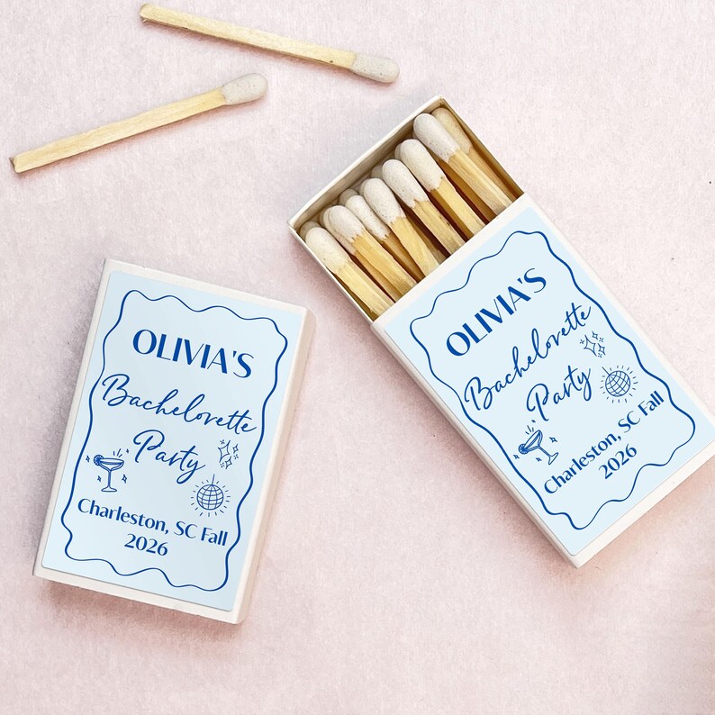 May include: Two white matchboxes with blue labels. The labels have a white background with blue text and illustrations. The text on the labels reads "Olivia's Bachelorette Party" and "Charleston, SC Fall 2026".