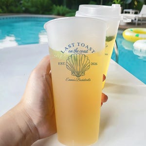 K&ouml;nnte beinhalten: Ein durchsichtiger Plastikbecher mit einem gelben Getr&auml;nk. Der Becher hat ein blaues und wei&szlig;es Design mit dem Text "Last Toast on the Coast", einer Muschelgrafik und dem Text "Est. 2026 Emma's Bachelorette".