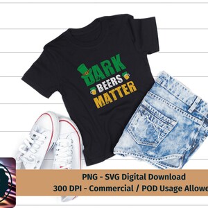 May include: A black t-shirt with a green shamrock and the text "Dark Beers Matter". The shirt is on a white wooden surface with a pair of white sneakers and a pair of blue jeans.