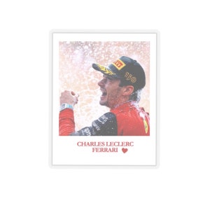 May include: A framed photograph featuring Charles Leclerc in a red Ferrari racing suit and cap, celebrating. The image includes the text "CHARLES LECLERC FERRARI" with a heart symbol. The background shows a celebratory spray.