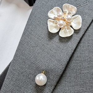 May include: A white flower brooch with pearl and rhinestone accents. The brooch is pinned to a gray suit jacket.