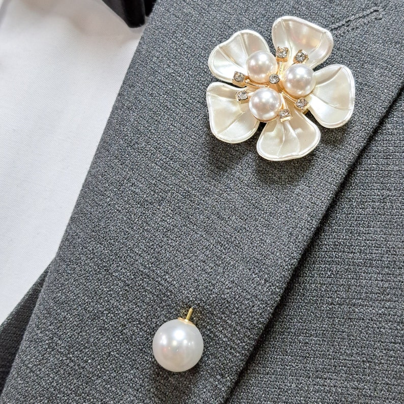 May include: A white flower brooch with pearl and rhinestone accents. The brooch is pinned to a gray suit jacket.