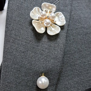 May include: A white flower brooch with pearl and rhinestone accents. The brooch is pinned to a gray fabric background..