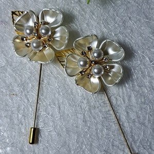 May include: Two white flower brooches with gold accents and pearl embellishments. Each brooch has a gold pin.