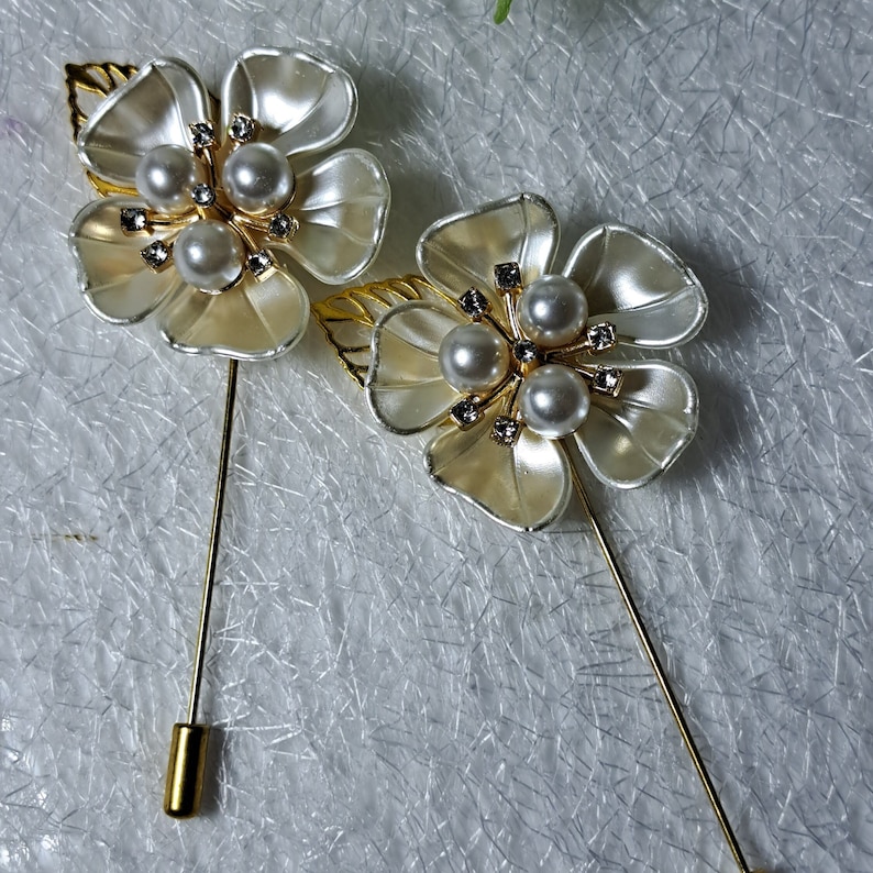 May include: Two white flower brooches with gold accents and pearl embellishments. Each brooch has a gold pin.