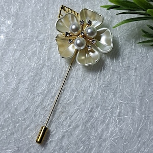 May include: A gold flower brooch with white petals and pearl accents. The brooch has a long, thin pin.