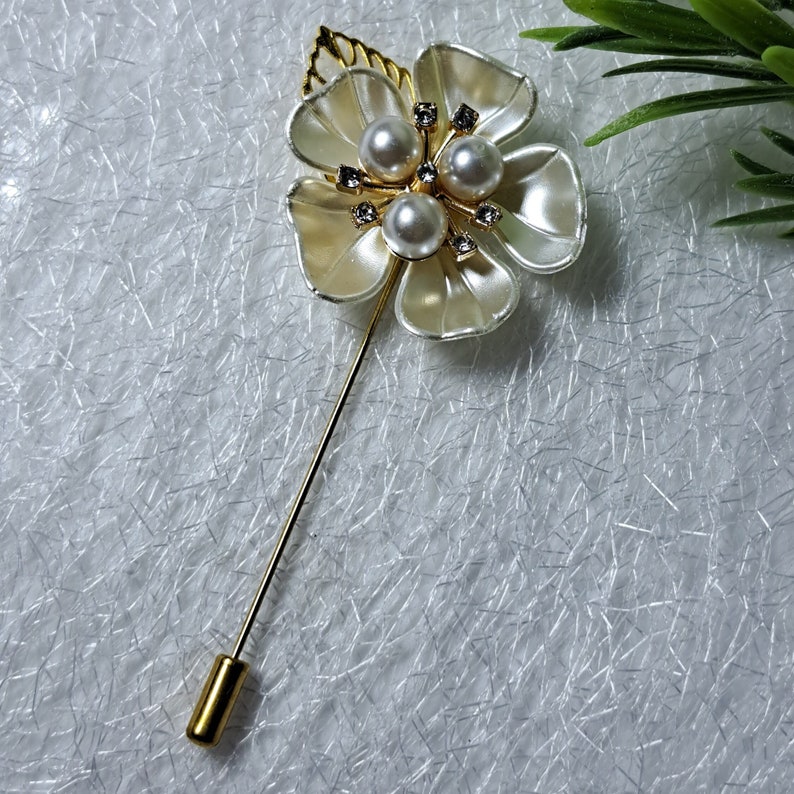 May include: A gold flower brooch with white petals and pearl accents. The brooch has a long, thin pin.