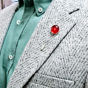 Puede incluir: Una chaqueta de tweed gris con una camisa de botones verde debajo. Un alfiler de vidrio rojo está sujeto a la solapa.