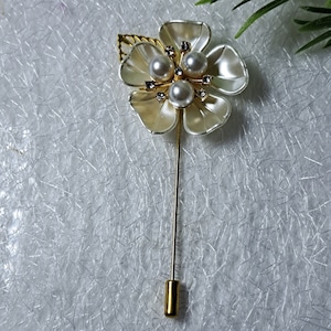 May include: A gold flower brooch with white petals and three faux pearls in the center. The brooch has a gold pin on the back.