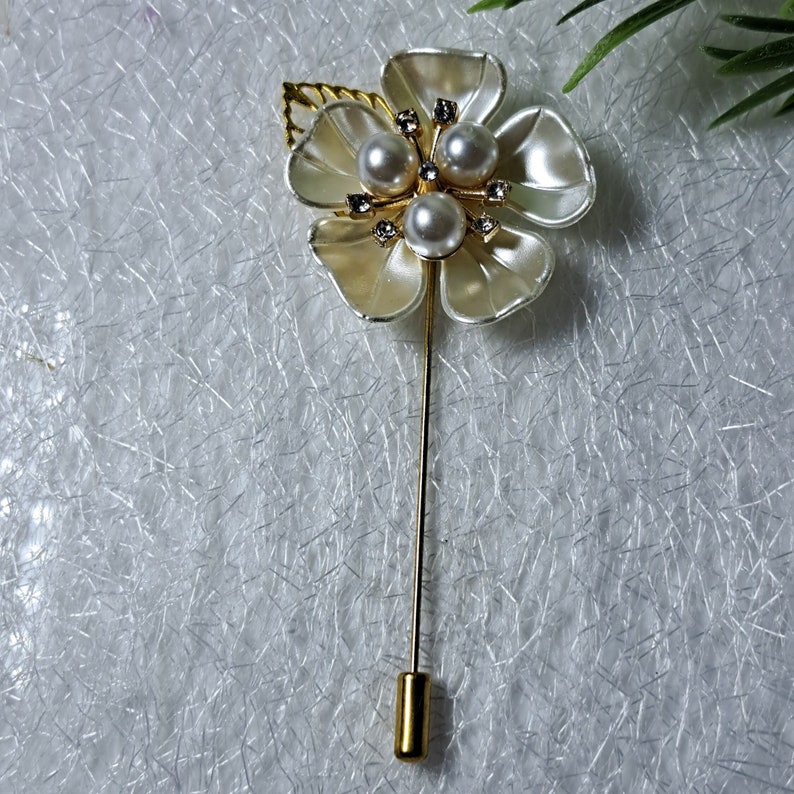 May include: A gold flower brooch with white petals and three faux pearls in the center. The brooch has a gold pin on the back.