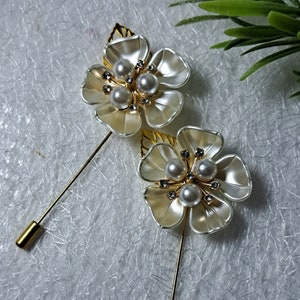 May include: Two gold brooches with white flower designs and pearl accents. Each brooch has a gold pin.