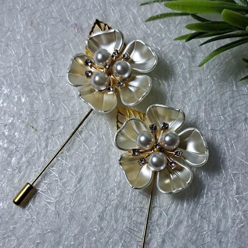 May include: Two gold brooches with white flower designs and pearl accents. Each brooch has a gold pin.
