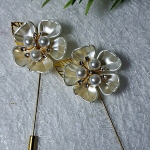 May include: Two gold tone flower brooches with white petals and pearl accents. Each brooch has a gold tone pin.