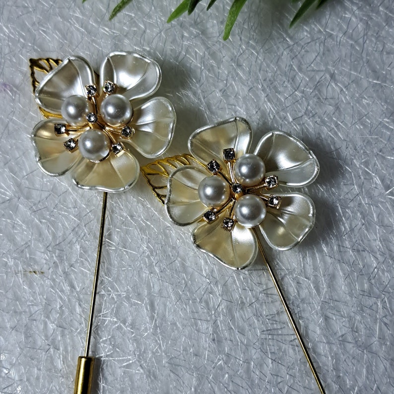 May include: Two gold tone flower brooches with white petals and pearl accents. Each brooch has a gold tone pin.