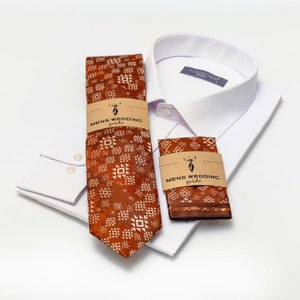 May include: A white dress shirt with a brown and white patterned tie and pocket square. The tie and pocket square have a brown background with white geometric designs. The tie and pocket square have a brown label with the text "Mens Wedding Guide" and a stylized deer head.