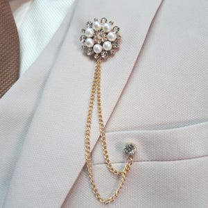 以下が含まれることがあります： 金色のブローチ。花柄のデザインで、イミテーションパールとクリアクリスタルが特徴です。繊細なゴールドチェーンがブローチから垂れ下がり、先端にはクリスタルが一つ。ブローチは薄いピンク色のブレザーに留められています。