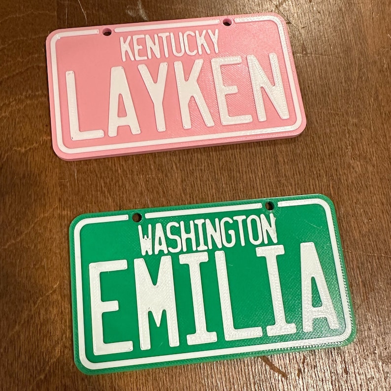 K&ouml;nnte beinhalten: Zwei personalisierte Schilder im Stil von Nummernschildern. Das obere Schild ist rosa mit wei&szlig;em Text, der "Kentucky Layken" lautet. Das untere Schild ist gr&uuml;n mit wei&szlig;em Text, der "Washington Emilia" lautet.