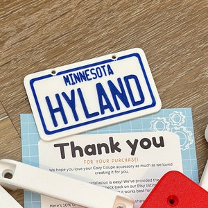 K&ouml;nnte beinhalten: Ein wei&szlig;es Plastikschild mit blauem Text, auf dem "MINNESOTA HYLAND" steht. Das Schild befindet sich auf einer Holzoberfl&auml;che mit einem Dankesschreiben und anderen Gegenst&auml;nden.