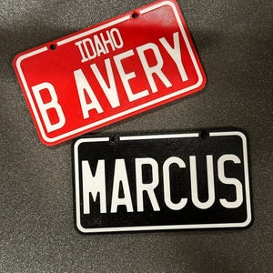 K&ouml;nnte beinhalten: Zwei personalisierte Schilder im Stil von Nummernschildern. Das obere Schild ist rot mit wei&szlig;en Buchstaben, auf denen "IDAHO B AVERY" steht. Das untere Schild ist schwarz mit wei&szlig;en Buchstaben, auf denen "MARCUS" steht.