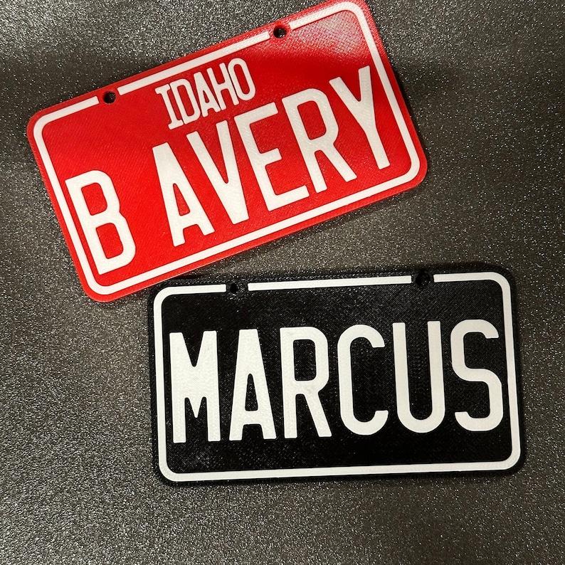 K&ouml;nnte beinhalten: Zwei personalisierte Schilder im Stil von Nummernschildern. Das obere Schild ist rot mit wei&szlig;en Buchstaben, auf denen "IDAHO B AVERY" steht. Das untere Schild ist schwarz mit wei&szlig;en Buchstaben, auf denen "MARCUS" steht.