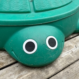May include: A green plastic turtle sandbox with large black eyes. The sandbox is sitting on a wooden deck.