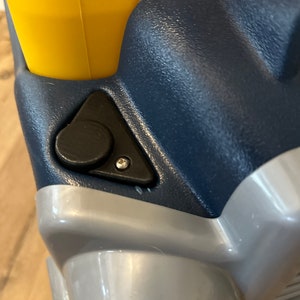 May include: Close-up of a blue and gray toy truck with a yellow handle. The truck has a black plastic button on the side.