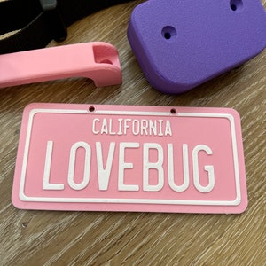 K&ouml;nnte beinhalten: Rosa Kunststoff-Kennzeichen mit wei&szlig;em Text "CALIFORNIA LOVEBUG".