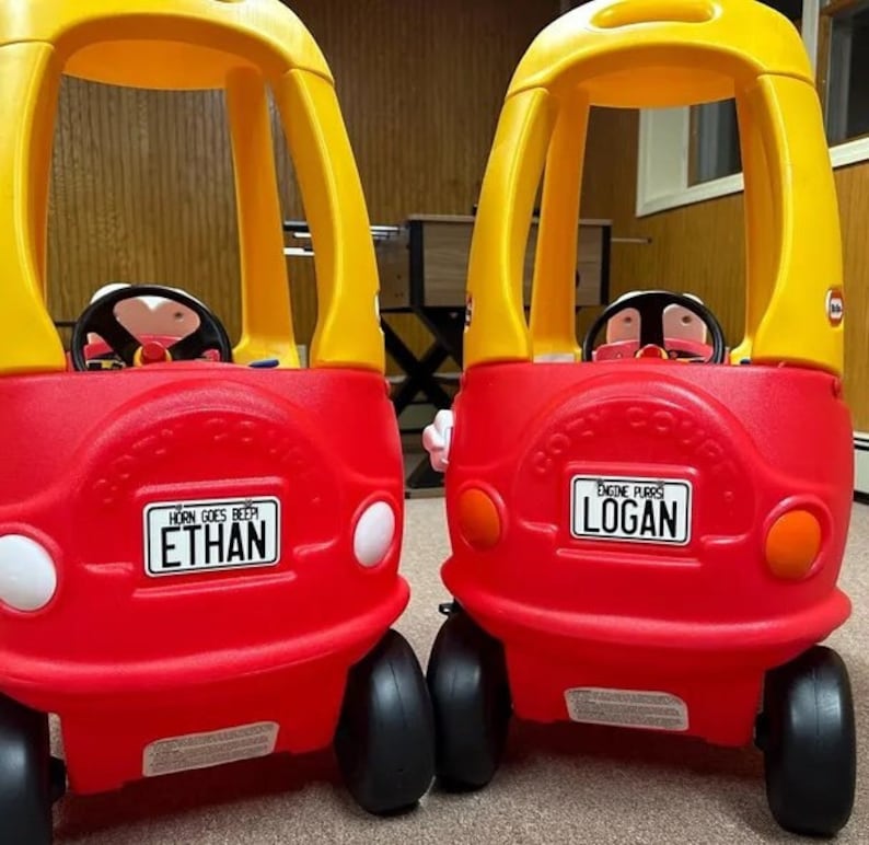 K&ouml;nnte beinhalten: Zwei rote und gelbe Spielzeugautos mit schwarzen R&auml;dern. Das Auto links hat ein Nummernschild mit der Aufschrift "Horn Goes Beep Ethan". Das Auto rechts hat ein Nummernschild mit der Aufschrift "Engine Purrs Logan".